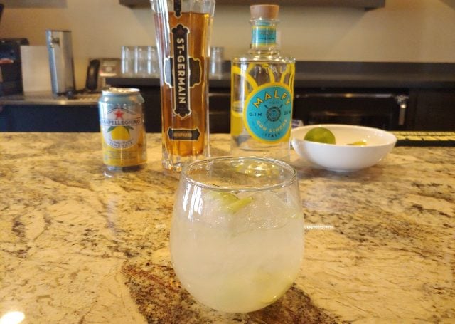 A cocktail placed in front of a can of lemonade, a St. Germain liquor bottle and a bottle of gin