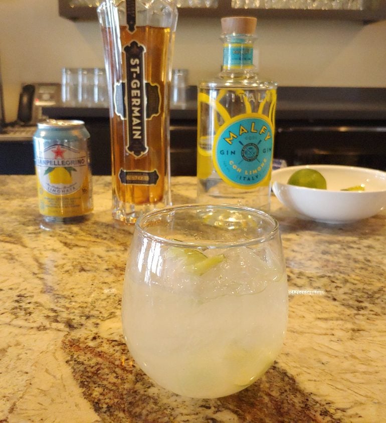 A cocktail placed in front of a can of lemonade, a St. Germain liquor bottle and a bottle of gin