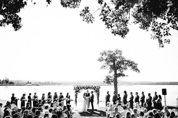 A black and white shot of guests looking on to the bride and groom at the aisle next to the bridesmaids and groomsmen.