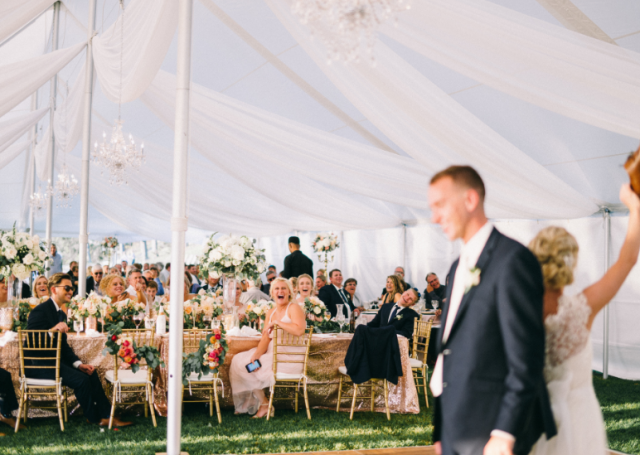 White tent wedding with guests seated on the dining table and looking on to the bride and groom