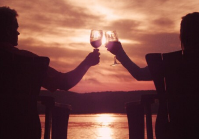 A couple clinking their wine glasses to cheers to the sunset by the water.