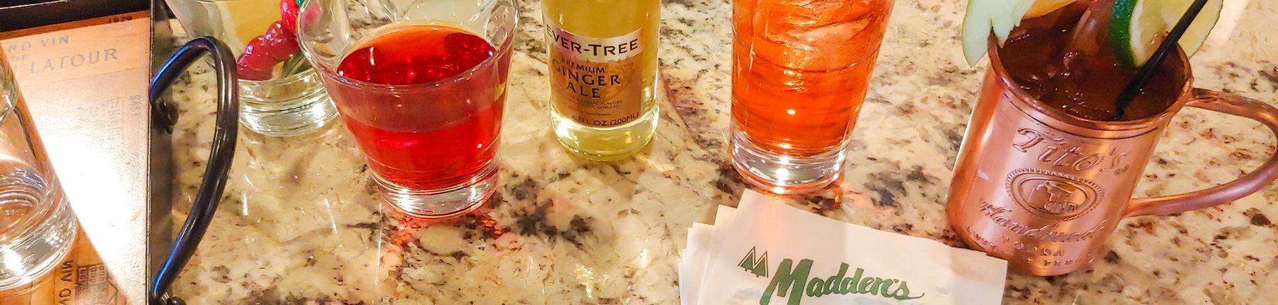 Cocktails next to a bottle of ginger ale placed on a marble counter with Madden's napkins